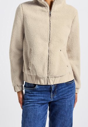 Veste sherpa beige zippée avec col haut et poignets élastiqués, portée avec un jean en denim bleu, vue de face à partir de la taille.