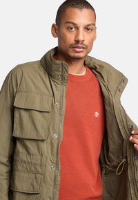 Chaqueta verde oliva con cuello alto, cierre de cremallera y múltiples bolsillos frontales, llevada sobre un suéter color óxido.