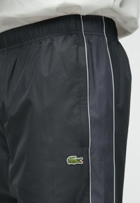 Μαύρα αθλητικά σορτς από ελαφρύ ύφασμα, με λευκή πλαϊνή ρίγα και κεντητό λογότυπο κροκόδειλου σε πράσινο χρώμα. Ελαστική ζώνη στη μέση.