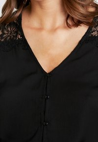 Blusa nera con scollatura a V, dettagli con bottoni e applicazioni in pizzo nero sulle spalle per aggiungere texture e design.