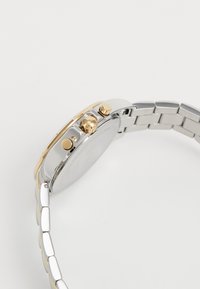 Montre en acier inoxydable argentée et dorée avec un boîtier rond, des cadrans chronographe et trois boutons-poussoirs sur le côté. Bracelet en métal lisse.