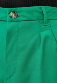 Gröna veckade byxor med en slät textur, som har en dekorativ knapp och bältesöglor, vilket framhäver en skräddarsydd design.