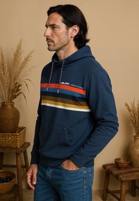 Marineblauwe hoodie met verstelbare trekkoorden, voorzien van opvallende horizontale strepen in rood, goud en wit. Zachte textuur, ontwerp met voorklep.