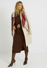 Un abrigo trench beige sobre un vestido de punto marrón con una abertura lateral, complementado por una bufanda de patrón burdeos y unos zapatos de tacón marrón.