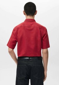 Rotes Kurzarmhemd mit Kragen und Aufhängeschlaufe aus strukturiertem Stoff. Kombiniert mit dunkler Jeanshose, von hinten betrachtet.