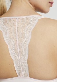 Lingerie van zachtroze stof, met ingewikkelde kantdetails aan de achterkant met golfpatronen en een geschulpt randje.