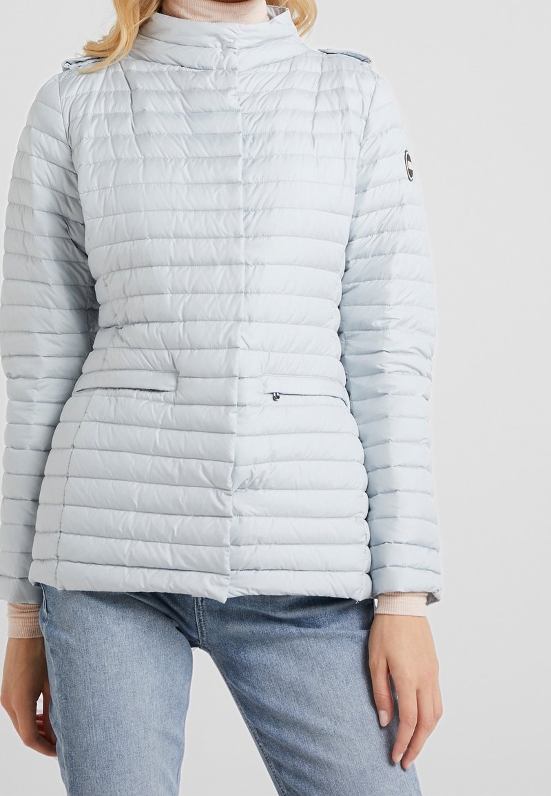 Světle modrá zateplená bunda s prošívaným vzorem, vysokým límcem a bočními kapsami na zip. Obsahuje tlačítka na ramenech a žebrované manžety.