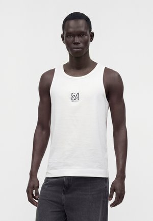 Homme portant un débardeur blanc sans manches avec un petit logo noir sur la poitrine et un pantalon gris foncé, debout devant un fond uni.