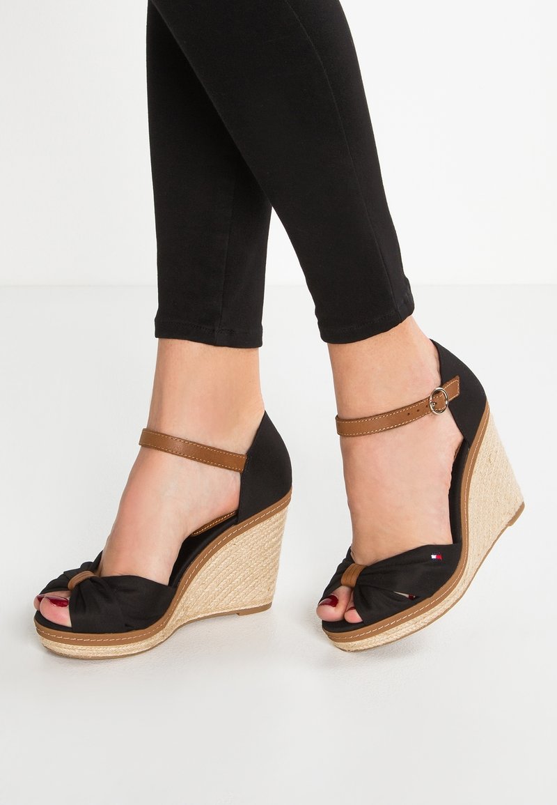 Pieds portant des sandales compensées noires avec des brides marron à la cheville et des talons tissés texturés, assorties à un pantalon noir coupé au-dessus de la cheville.