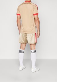 Beige kortärmad skjorta med röda accenter, tillsammans med glänsande beiga shorts med röda sidoband och vita knälånga strumpor med svarta ränder.