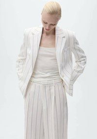 Bílý pruhovaný blejzr se sladěnými kalhotami, vyznačující se tailored designem a lehkým materiálem. Doplněno o přiléhavý bílý top.
