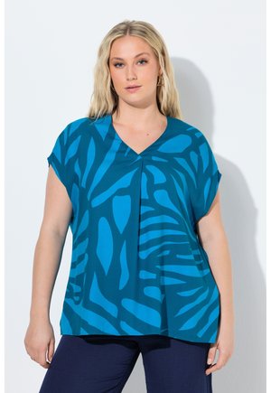 Frau mit langen blonden Haaren, die eine blaue, gemusterte V-Ausschnitt-Bluse und dunkle Hosen trägt, steht vor einem weißen Hintergrund.