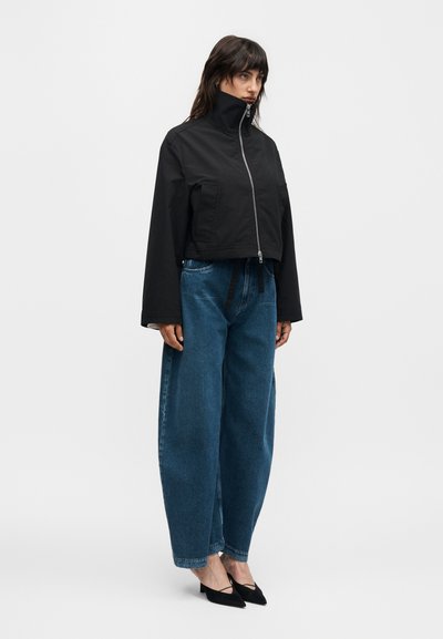 Γυναίκα που φοράει μαύρο μπουφάν με φερμουάρ και ψηλό γιακά, φαρδιά μπλε τζιν και μαύρες μυτερές γόβες, στέκεται μπροστά σε λευκό φόντο.