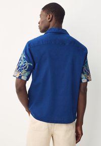 Chemise à manches courtes en tissu bleu vif ; présente une broderie florale colorée sur les manches et un col classique.