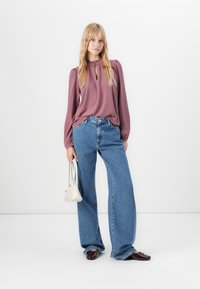 Donna che indossa una blusa a maniche lunghe color malva e jeans a gamba larga blu, mentre tiene una piccola borsa bianca, in piedi contro uno sfondo bianco uniforme.