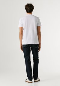 Kiharatukkainen mies, joka pukeutunut yksiväriseen valkoiseen lyhythihaiseen t-paitaan, tummiin farkkuihin ja valkoisiin tennareihin, seisoo selin yksinkertaista taustaa vasten.