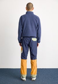 Pull en polaire marine avec un col montant, associé à un pantalon marine avec des blocs de couleur orange et verte et des accents en patch.