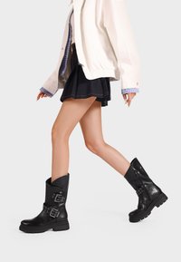 Stivali alla caviglia in pelle nera con suola spessa, cerniera laterale e dettagli decorativi in fibbia e borchie. Texture liscia con bordo ripiegato.