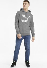 Szürke kapucnis pulóver, nagy fehér Puma logóval, elülső zsebbel, bordázott ujjak és alsó szegély, kék farmernadrággal és fekete sportcipővel párosítva.