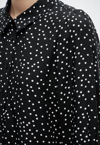 Μαύρο πουκάμισο με κουμπιά και άσπρες ακανόνιστες πουά, με μυτερό γιακά και μακριά μανίκια σε ένα άτομο.