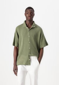 Camicia a maniche corte, con bottoni, realizzata in tessuto di maglia verde oliva testurizzato. Presenta un colletto ampio e una vestibilità rilassata, abbinata a pantaloni chiari.