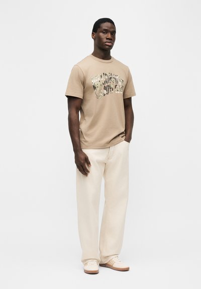 Νεαρός άνδρας που φοράει ρούχο τύπου t-shirt χρώματος μπεζ με σχέδιο, παντελόνι εκρού και λευκά αθλητικά, στέκεται με το ένα χέρι στην τσέπη μπροστά σε ένα απλό λευκό φόντο.