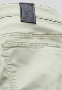 Lichtgroene broek met een leren label waarop "M5" staat. Heeft zichtbare stiksels en twee achterzakken. Gladde stofstructuur.
