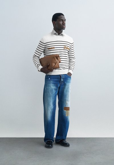 Homme portant un pull à rayures blanches et noires, jean bleu avec des patchs, chaussures noires, tenant un sac en cuir marron plié, debout contre un mur gris uni.