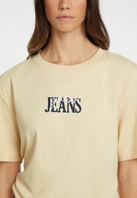 Jasnożółta bawełniana koszulka z krótkim rękawem, z granatowym i białym logo graficznym z napisem "Guess Jeans". Okrągły dekolt.