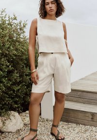 Top court en lin beige avec des liens sur le côté, associé à un short plissé beige clair. Le modèle porte des sandales vertes avec des éléments décoratifs.