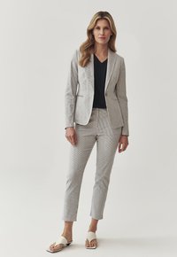 Gestreifter hellgrauer Blazer mit passenden maßgeschneiderten Hosen und einem schwarzen V-Ausschnitt-Oberteil, ergänzt durch weiße Slip-On-Offene-Schuhe.