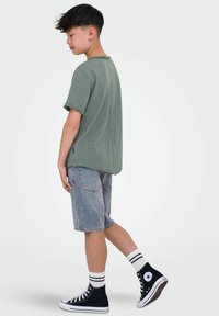Zielona koszulka z krótkim rękawem o luźnym kroju, w zestawie z jasnoszarymi szortami dżinsowymi i granatowymi trampkami za kostkę. Widoczne białe skarpetki w paski.