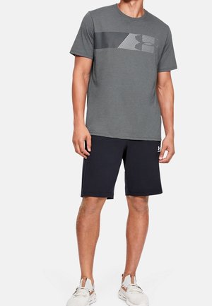 Mann trägt ein graues T-Shirt mit Grafik, schwarze Sportshorts und weiße Sneakers, steht vor einem schlichten hellen Hintergrund.