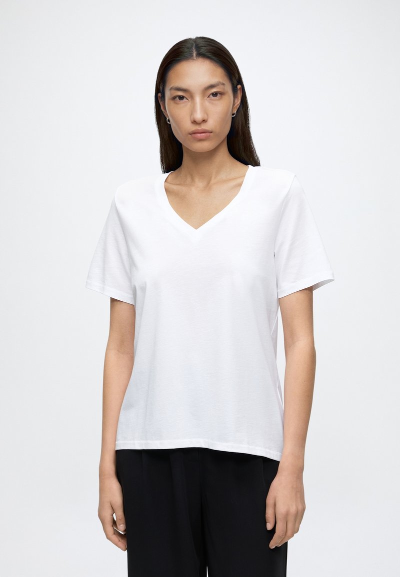 Giovane donna con lunghi capelli scuri indossa una semplice T-shirt bianca con scollo a V e pantaloni neri su uno sfondo grigio chiaro uniforme.
