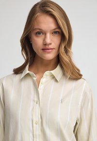 Camicia button-up color crema con righe verticali azzurro chiaro, dotata di un logo ricamato discreto sul lato sinistro del petto e di un colletto classico.