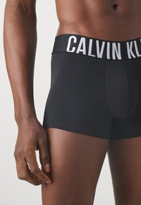 Melnas kokvilnas bokseršorti ar kontrastējošu baltu jostasvītru, uz kuras redzams "CALVIN KLEIN" logotips. Gluda tekstūra, cieša piegriezuma un vidēja augstuma dizains.