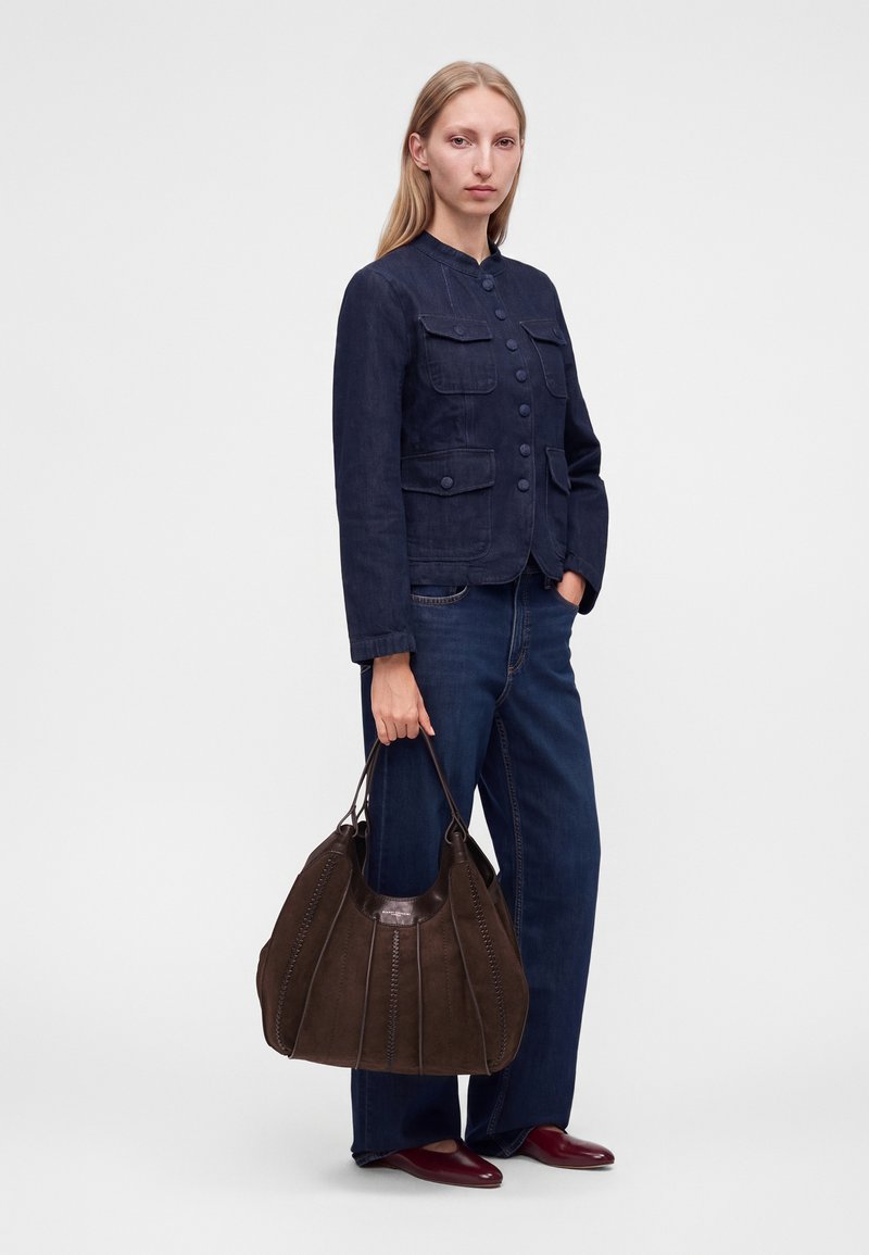 Femme aux longs cheveux blonds portant une veste bleu foncé boutonnée, un jean bleu, des chaussures bordeaux, tenant un grand sac à main marron à côté d'elle.