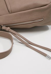 Angolo di una borsa in pelle taupe con zip argentata visibile e due lunghe nappine in pelle accanto a una tracolla coordinata su una superficie bianca.