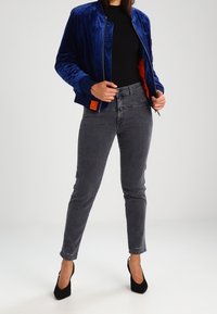 Woman wearing a blue velvet bomber jacket over a black top, gray high-waisted jeans, and black high-heeled shoes standing against a white background.