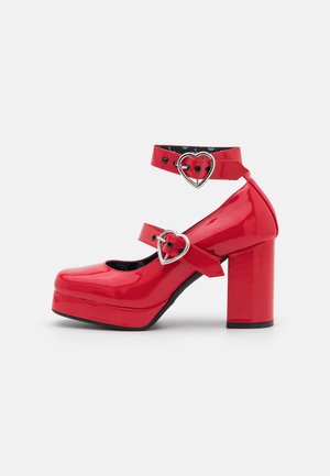 Talons à plateforme en cuir verni rouge avec des brides à la cheville et au médio-pied maintenues par des boucles argentées en forme de cœur.