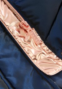 Gros plan sur une tirette de fermeture éclair en métal or rose sur un tissu bleu marine avec une bordure brillante or rose, embossée du mot Baker.