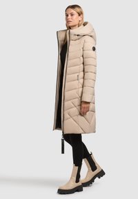 Bež puhovka z visokim ovratnikom, prešita zasnova, stranski žepi in srebrnim zadrgo. Kombinirana s kremnimi gležnarji in črnimi pajkicami.