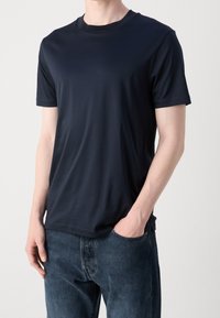 Man som bär en enkel svart kortärmad t-shirt med blåjeans, med en hand i fickan, stående mot en neutral bakgrund.