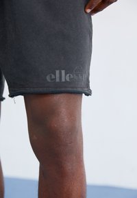 Svarte shorts med frynser i kanten, med en teksturert finish og "Ellesse"-logoen i en litt lysere print på venstre lår.