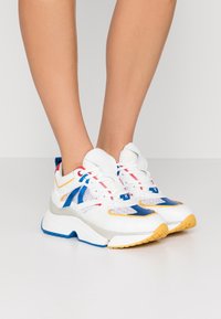 Person som bär vita sneakers med blå, röda och gula accenter och kraftiga sulor, stående på ett vitt golv mot en enkel bakgrund.