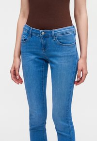 Γυναίκα που φοράει ανοιχτό μπλε skinny τζιν και καφέ τοπ, στέκεται με τα χέρια χαλαρά πλάι στο σώμα της, μπροστά σε απλό φόντο.