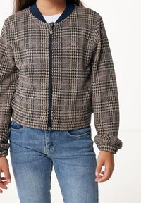 Hnědo-modrá bomber bunda s kostkovaným vzorem, se zipem vpředu, žebrovaným límcem a elastickými manžetami. Nositá přes bílou košili a modré džíny.