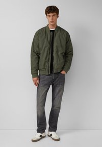 Olivgrüne Bomberjacke mit Reißverschluss, gerippte Bündchen, getragen über einem schwarzen Shirt, kombiniert mit verwaschenen Jeans und weißen Sneakern.