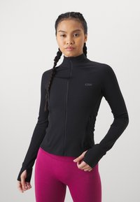 Melna sporta jaka ar rāvējslēdzēju, garām piedurknēm, stiepjamā audumā un sānu kabatām, uzvilkta virs rozā pieguļošām legingām. Jaka ar logotipu.