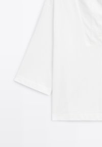 Chemise blanche à manches courtes avec une poche poitrine visible, montrée de la moitié du corps à la manche gauche.